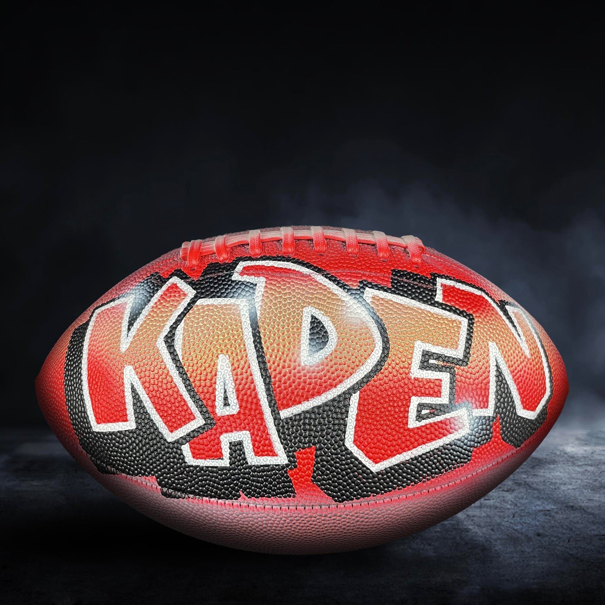 A football with the name "Kaden" painted on it, against a dark background with smoke.