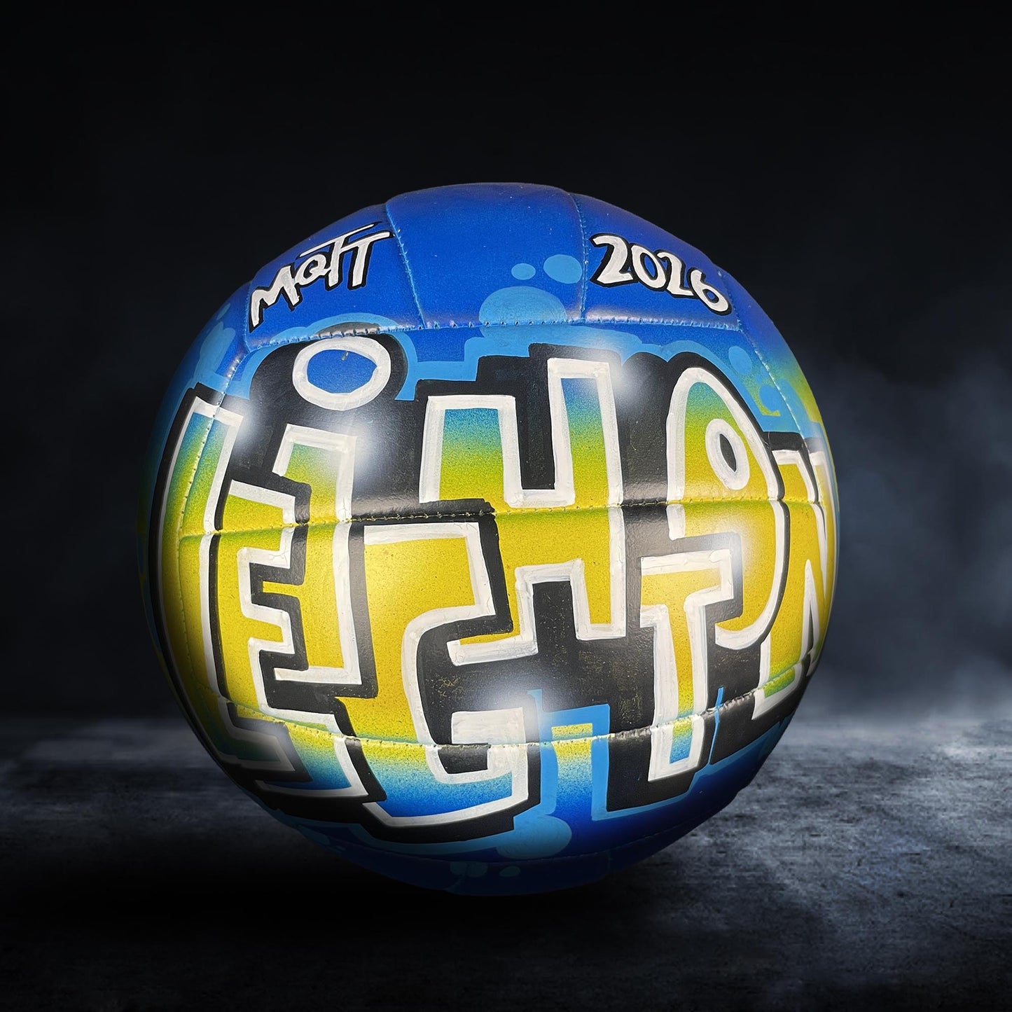A blue and yellow volleyball with the word "MATT" and the year "2016" written on it, against a dark background.