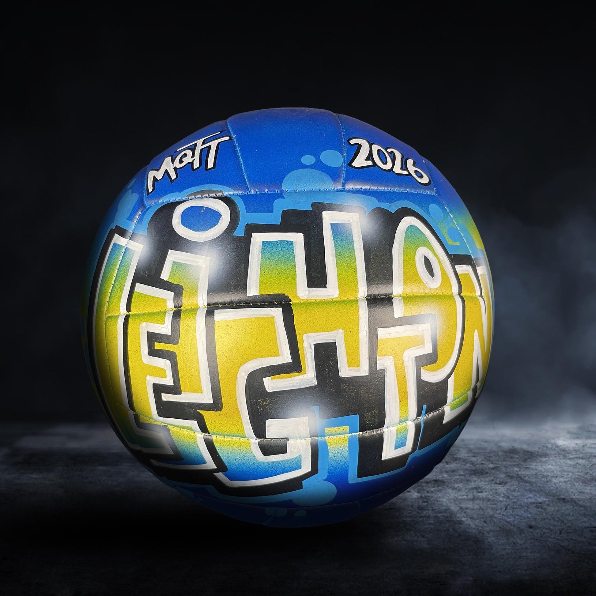 A blue and yellow volleyball with the word "MATT" and the year "2016" written on it, against a dark background.