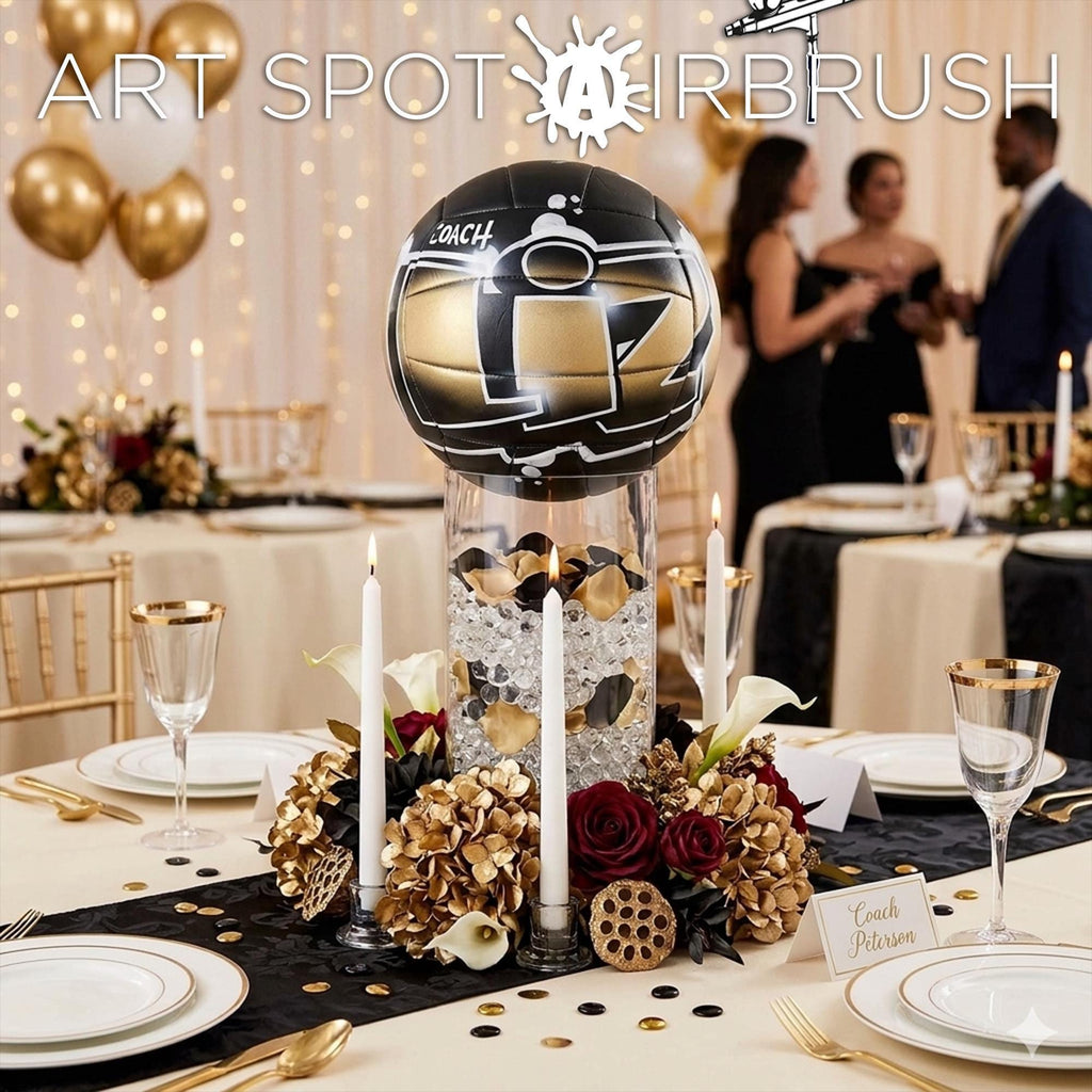 The image is a table setting for a formal event, featuring a centerpiece of a black and gold sports ball surrounded by gold and red flowers, candles, and confetti. The table is set with white plates, gold and silver cutlery, and champagne glasses. In the background, there are people dressed in formal attire, suggesting a social gathering or celebration.