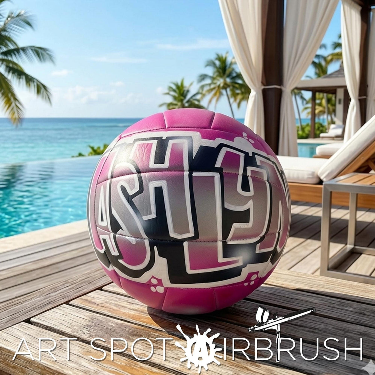 A pink and black custom volleyball personalized with the name "ASHLYN" written on it, sitting on a wooden deck near a pool and palm trees.