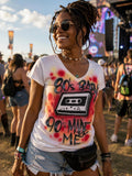 A smiling woman wearing a white t-shirt with a graphic design, sunglasses, and a necklace is standing in a crowd at what appears to be a music festival.