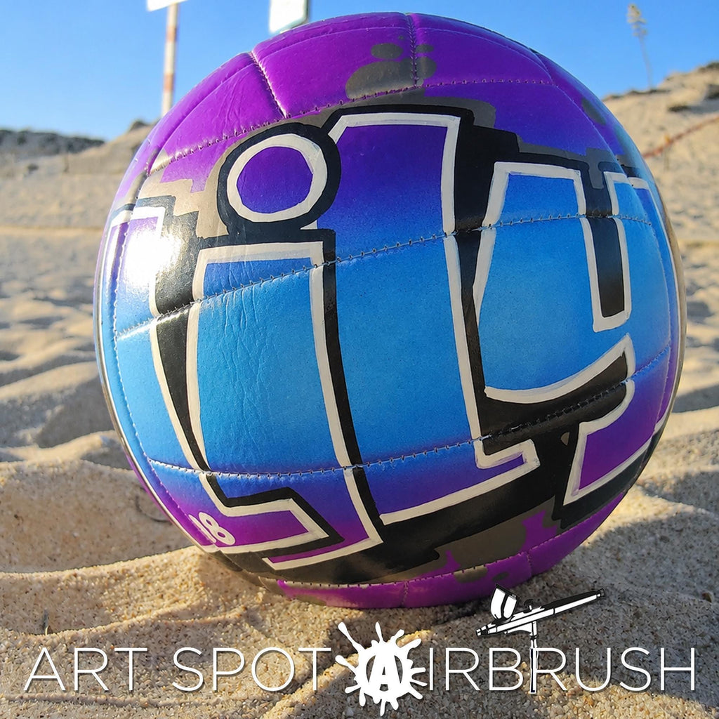 A purple and blue custom volleyball personalized with graffiti-style lettering on it, resting on a sandy beach.