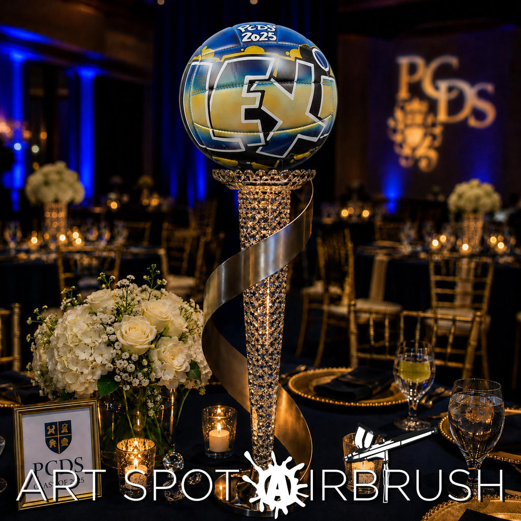 A table setting with a graffiti-style custom volleyball sitting on a large, ornate trophy-shaped centerpiece, surrounded by white flowers and candles. The table is set for a formal event, with a large screen displaying the letters "PCDS" in the background.