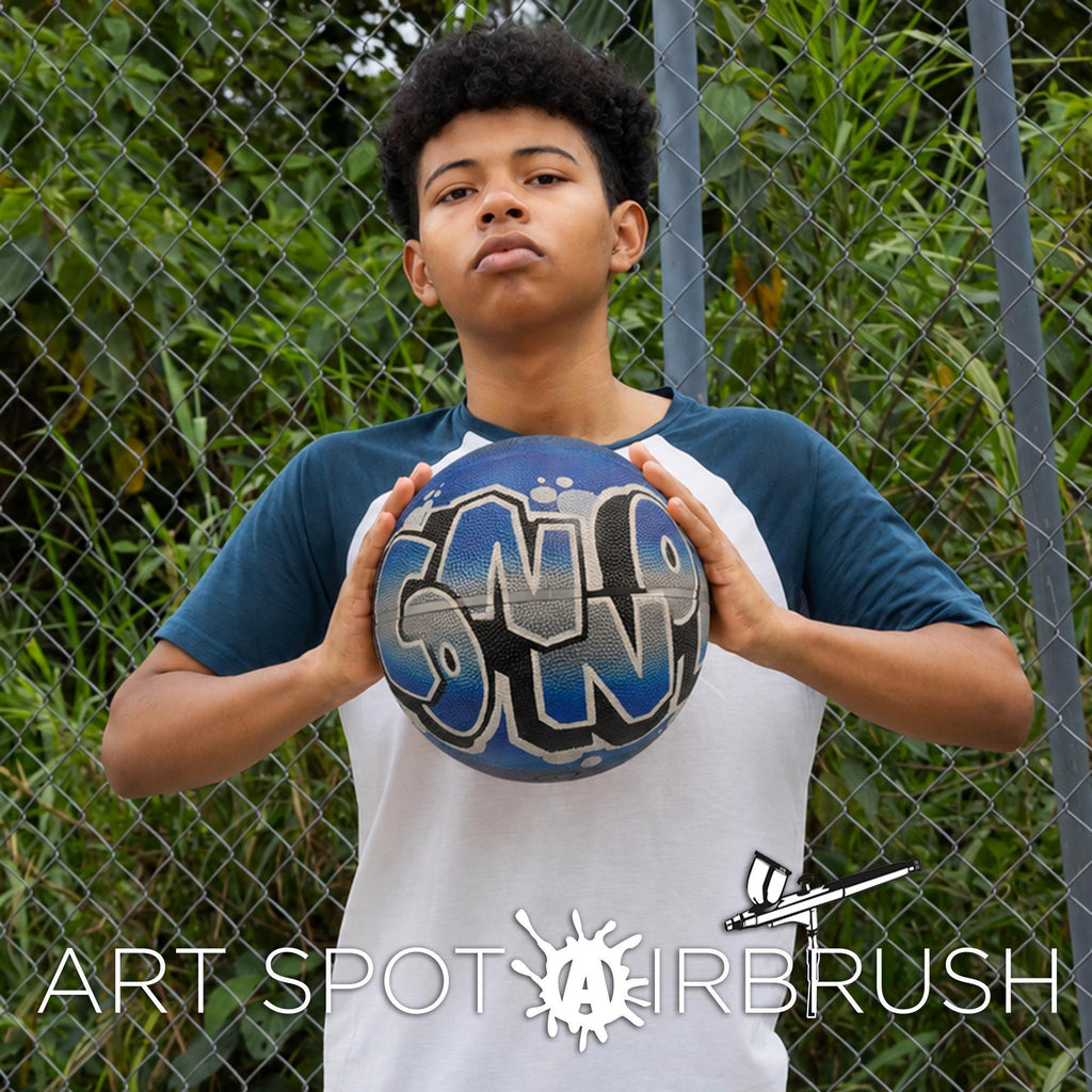 A young man holding a soccer ball with graffiti on it, standing in front of a chain-link fence.