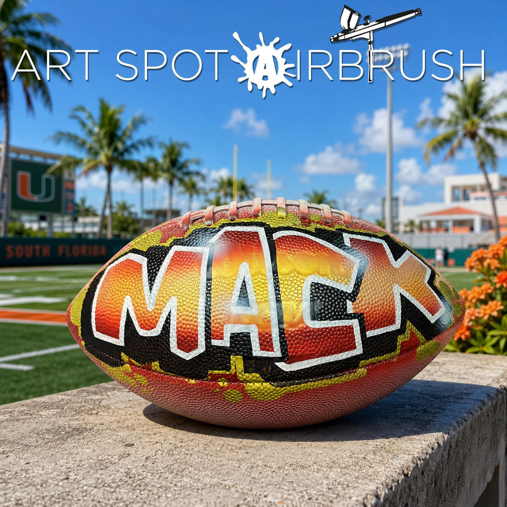 A custom football personalized with the name "MACK" painted on it, sitting on a concrete ledge with palm trees and a clear blue sky in the background.