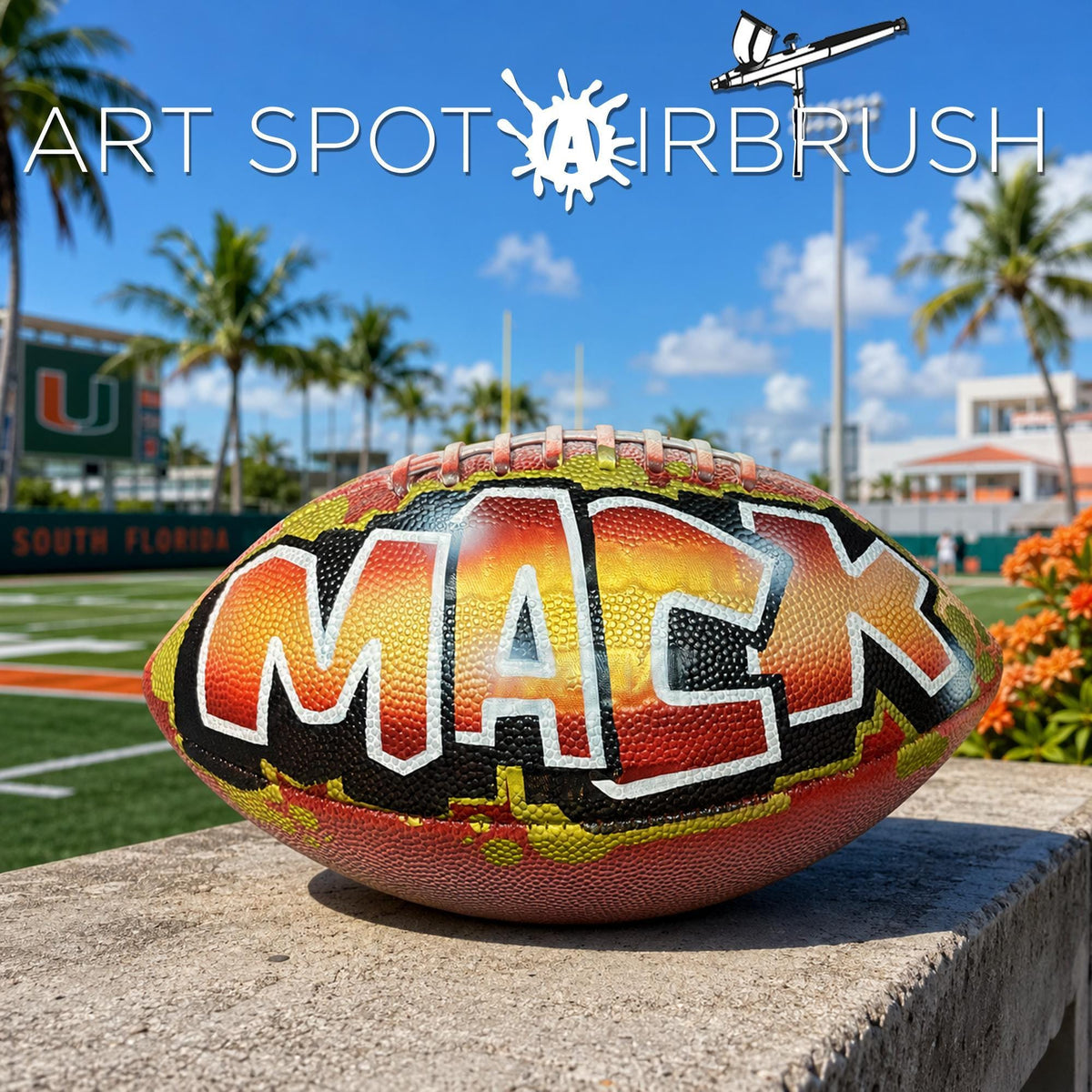 A custom football personalized with the name "MACK" painted on it, sitting on a concrete ledge with palm trees and a clear blue sky in the background.