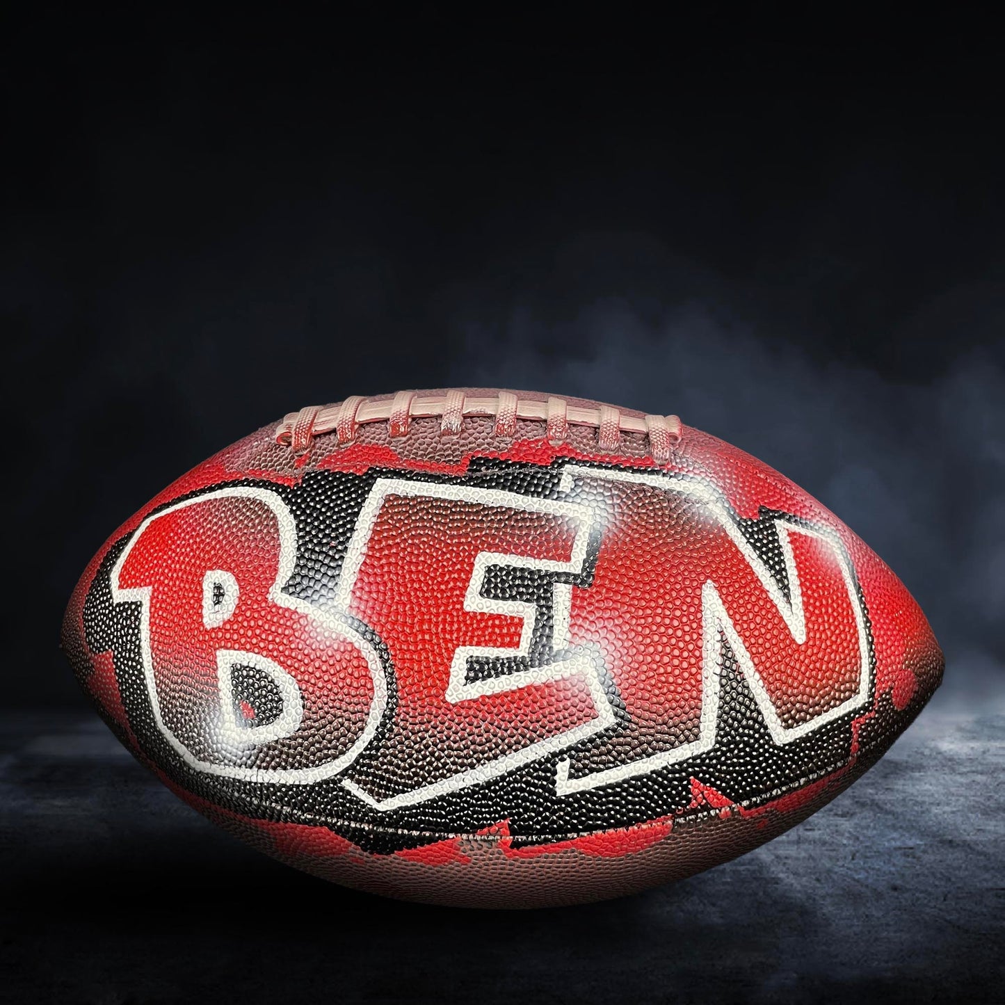 A red and black football with the name "BEN" painted on it, sitting on a dark surface with smoke or fog in the background.