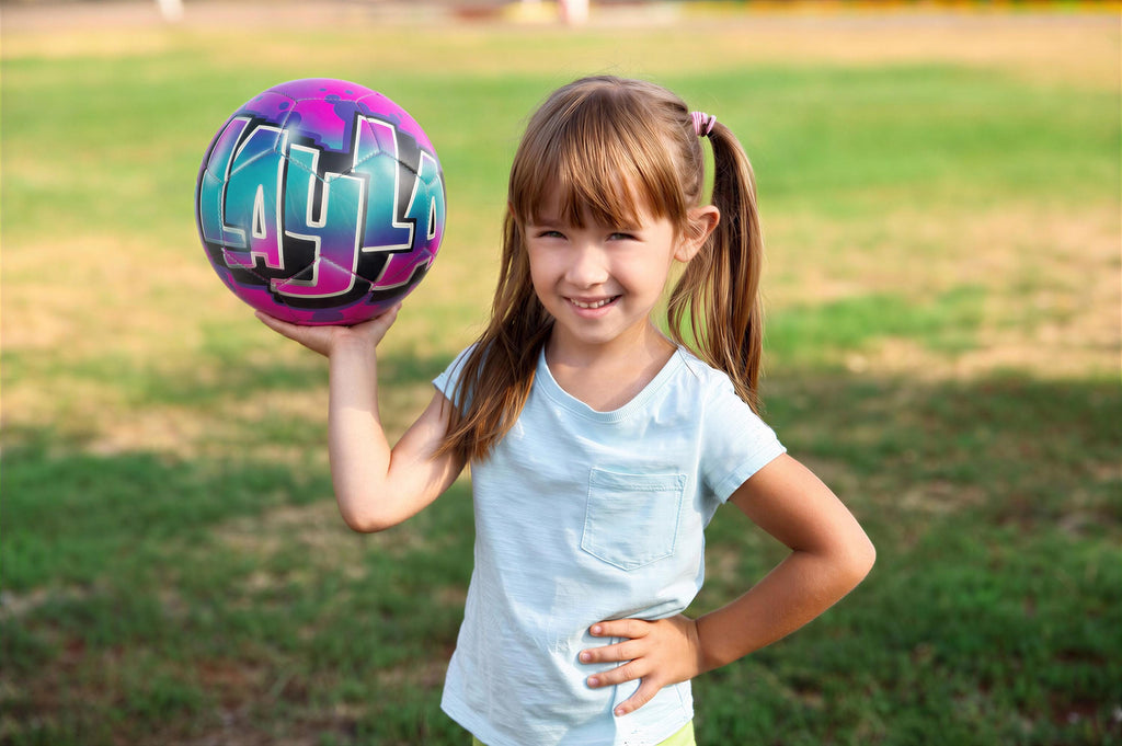 A young girl is holding a colorful sports ball with the word "LAYLA" written on it, smiling and posing in a grassy field.