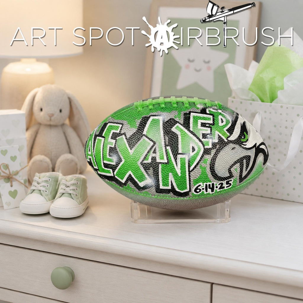 a personalized football with the name "ALEXANDER" painted on it, placed on a white dresser next to a pair of baby shoes.
