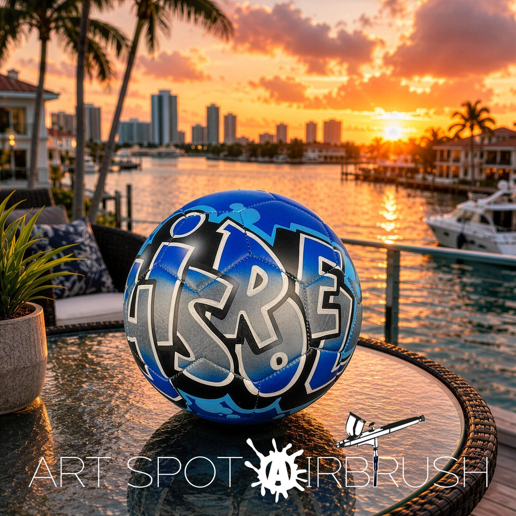 A blue and black soccer ball with the word "ISRE" written on it, placed on a table near a body of water with palm trees and a city skyline in the background during a sunset.