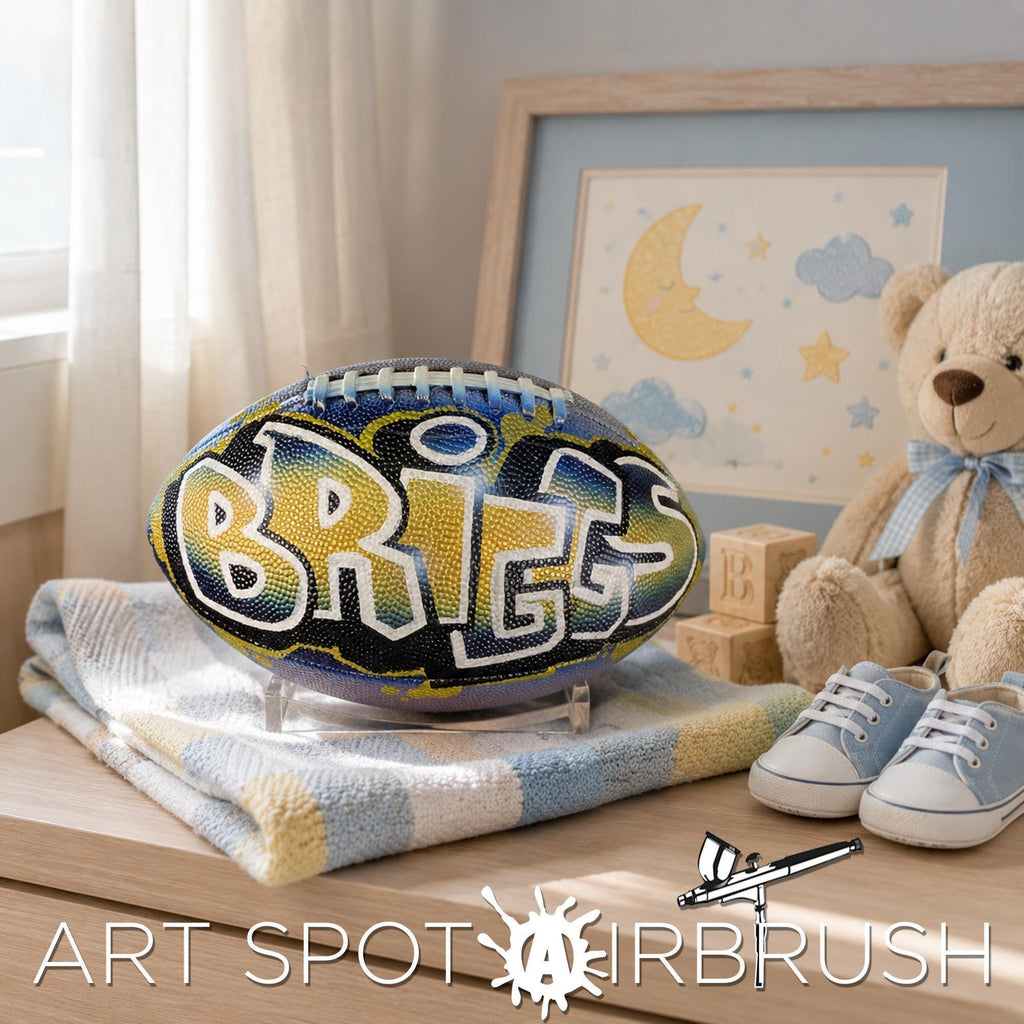 A personalized football with the name "BRIGGS" painted on it, placed on a wooden surface next to a teddy bear and baby shoes.