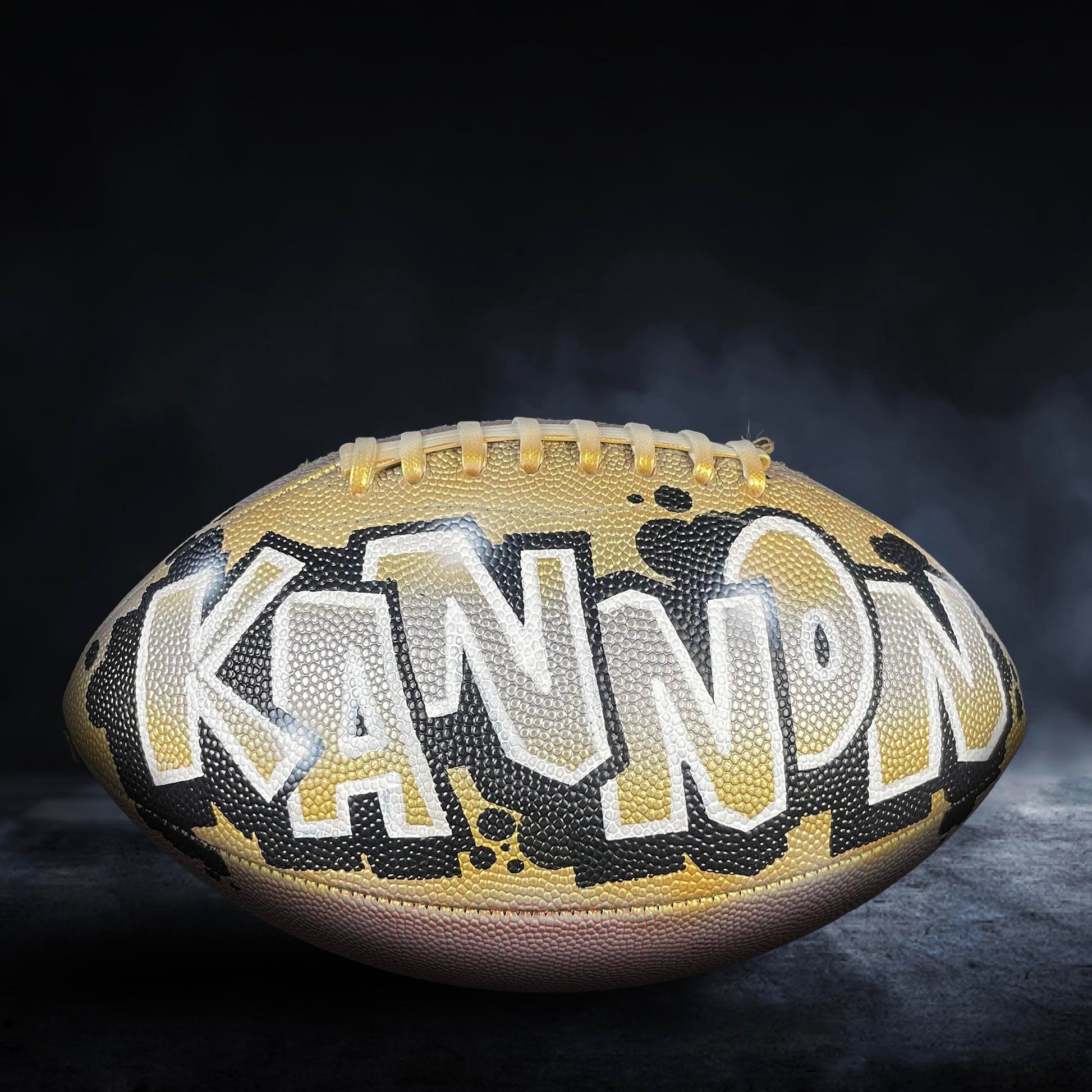 A football with the word "KANNON" painted on it, against a dark background with smoke or fog.