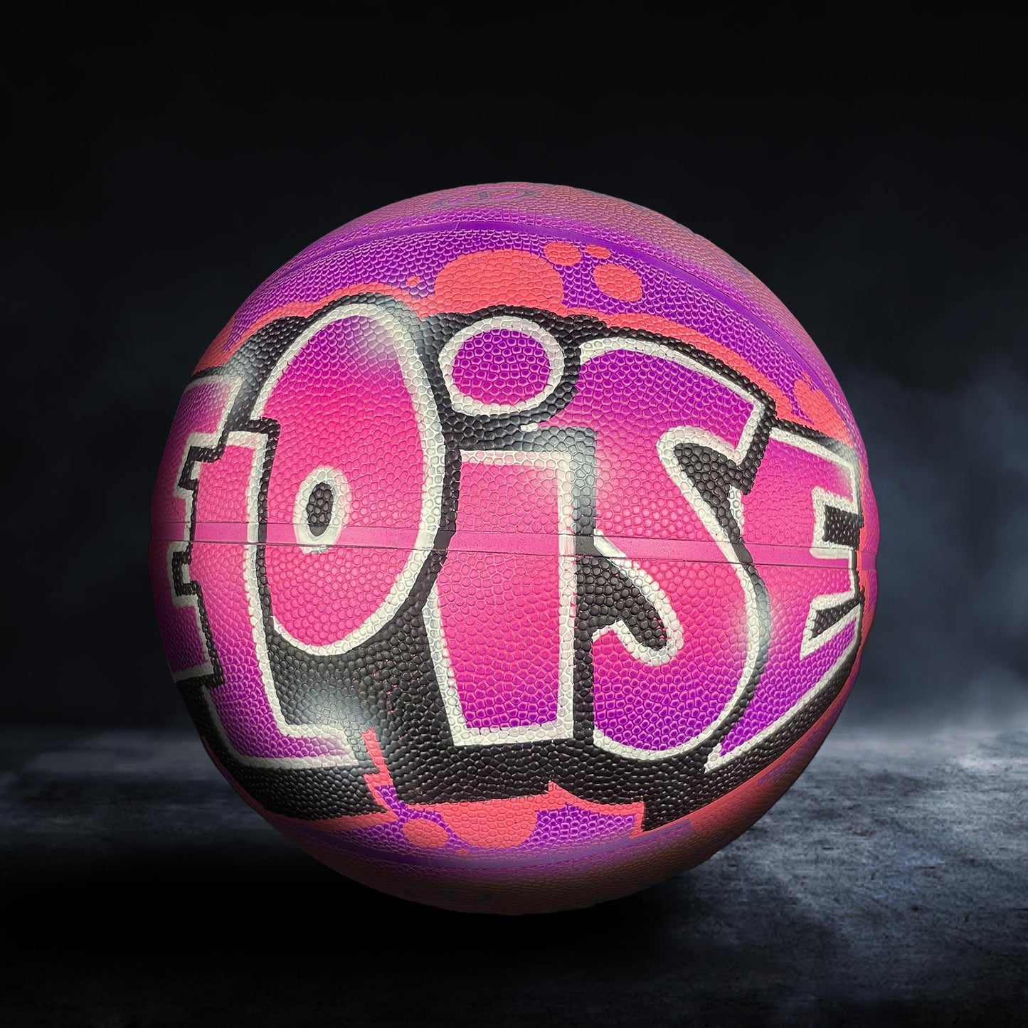 A colorful basketball with the word "OISE" painted on it, set against a dark background.