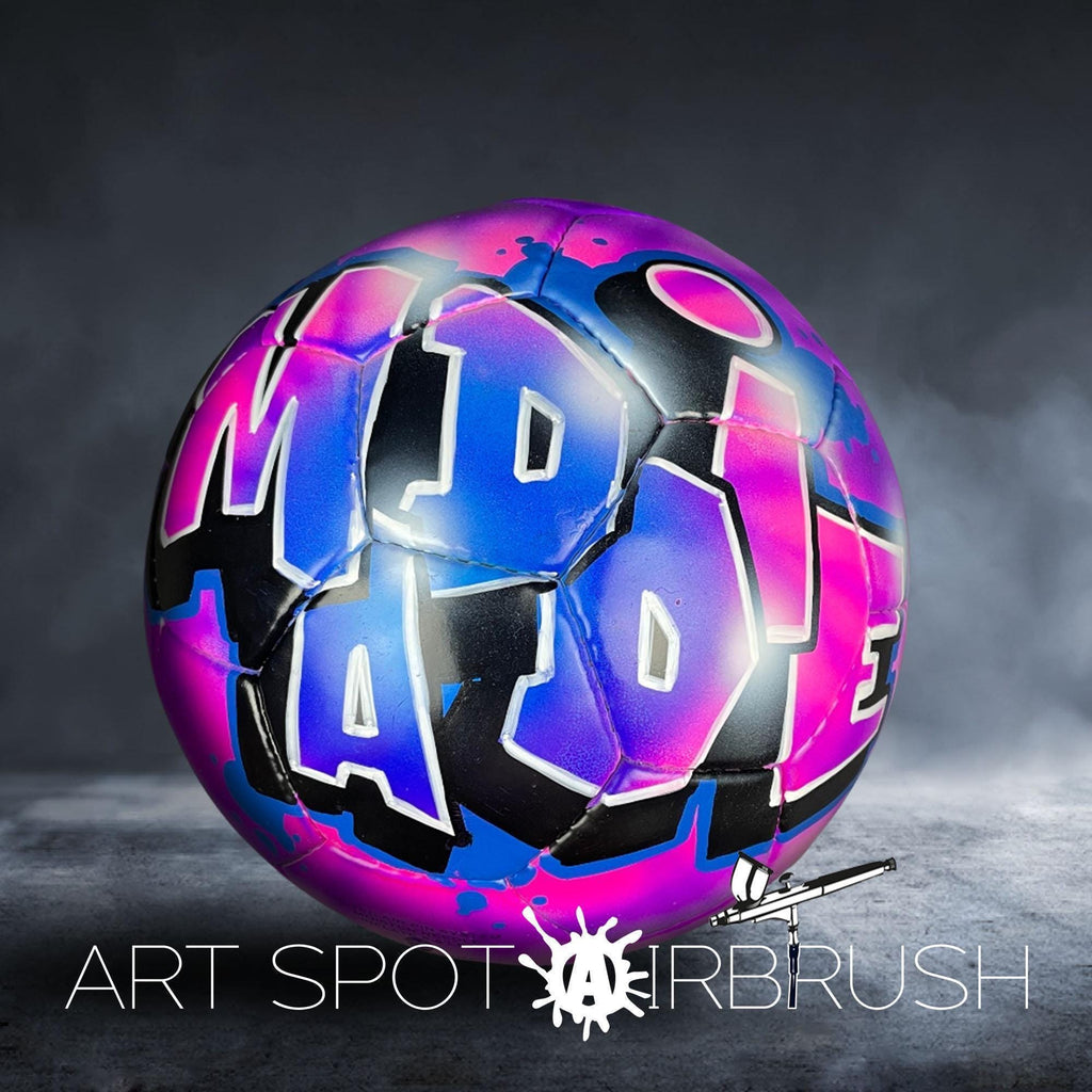 A colorful, graffiti-style soccer ball with the word "MAD" painted on it, set against a dark background with smoke or fog.