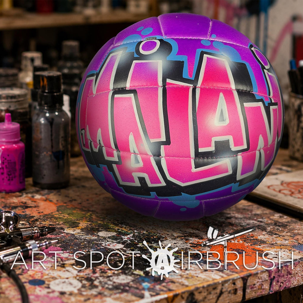 A colorful, graffiti-style volleyball with the word "MILAN" painted on it, sitting on a cluttered work surface with various art supplies and equipment in the background.