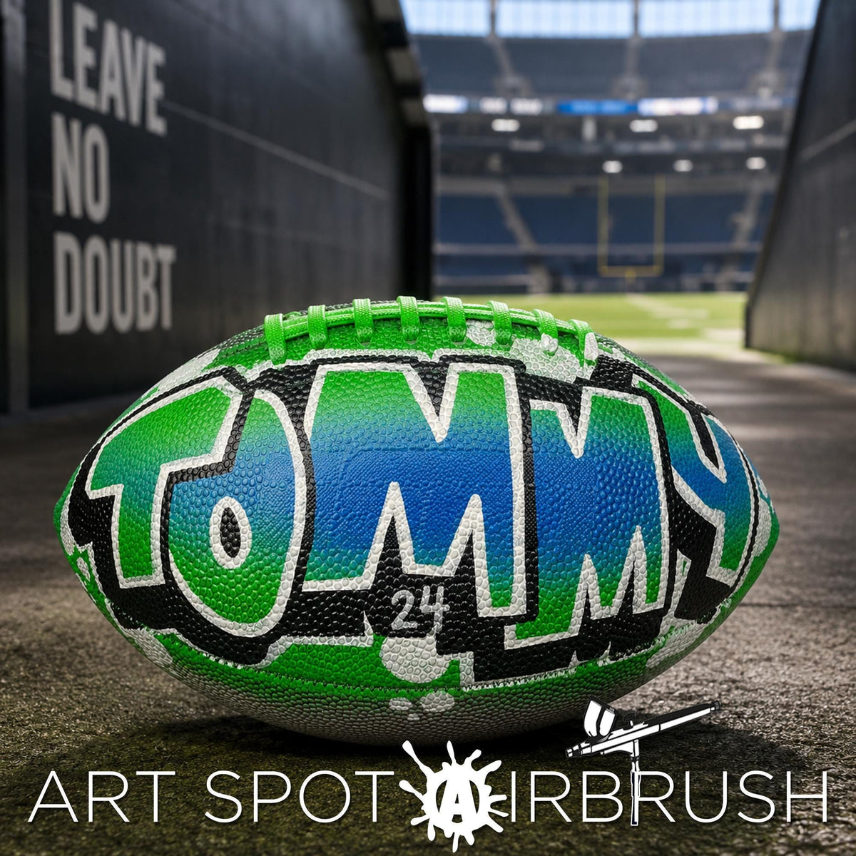 A personalized football with the name "TOMMY" painted on it, sitting on a concrete surface in an indoor stadium setting.