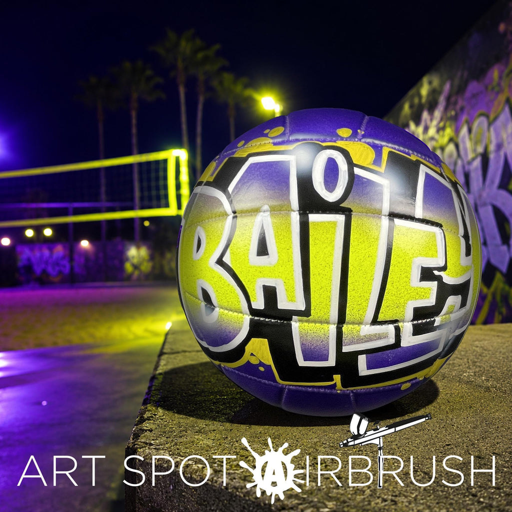 A purple and yellow custom volleyball personalized with the word "BAILEY" painted on it, sitting on a concrete surface at night with a graffiti-covered wall in the background.