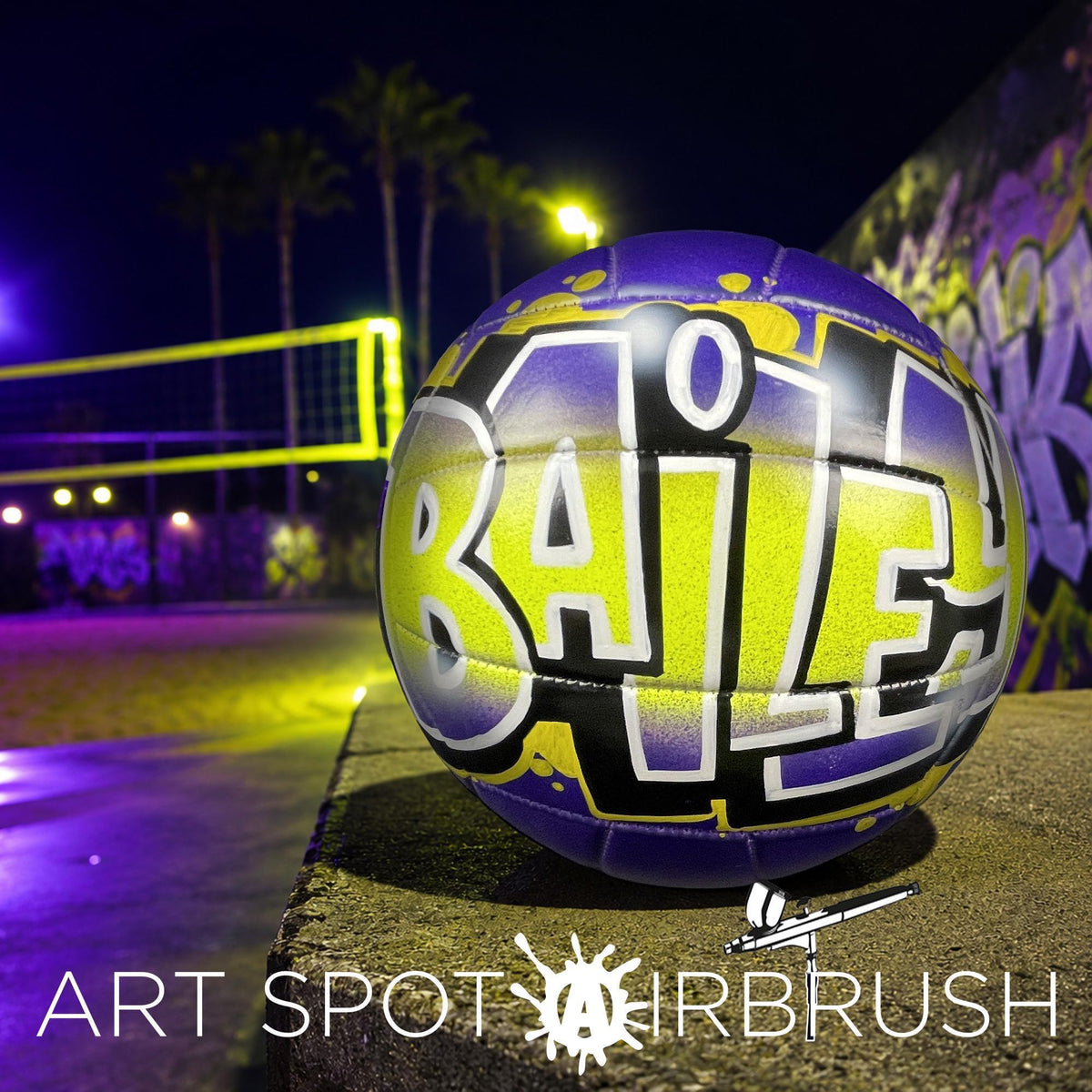 A purple and yellow custom volleyball personalized with the word "BAILEY" painted on it, sitting on a concrete surface at night with a graffiti-covered wall in the background.