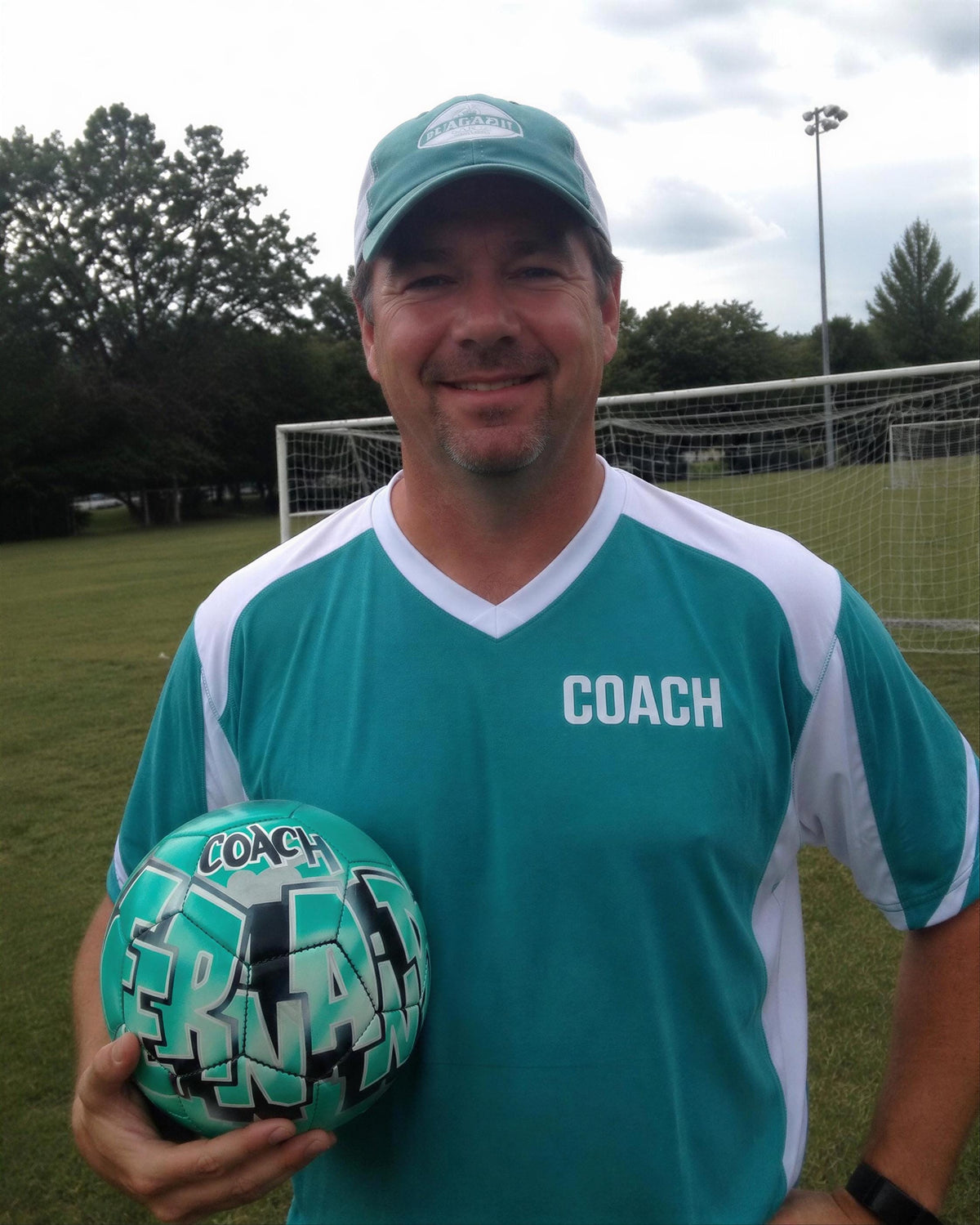 A man in a green and white soccer jersey is holding a soccer ball with the word "COACH" written on it.