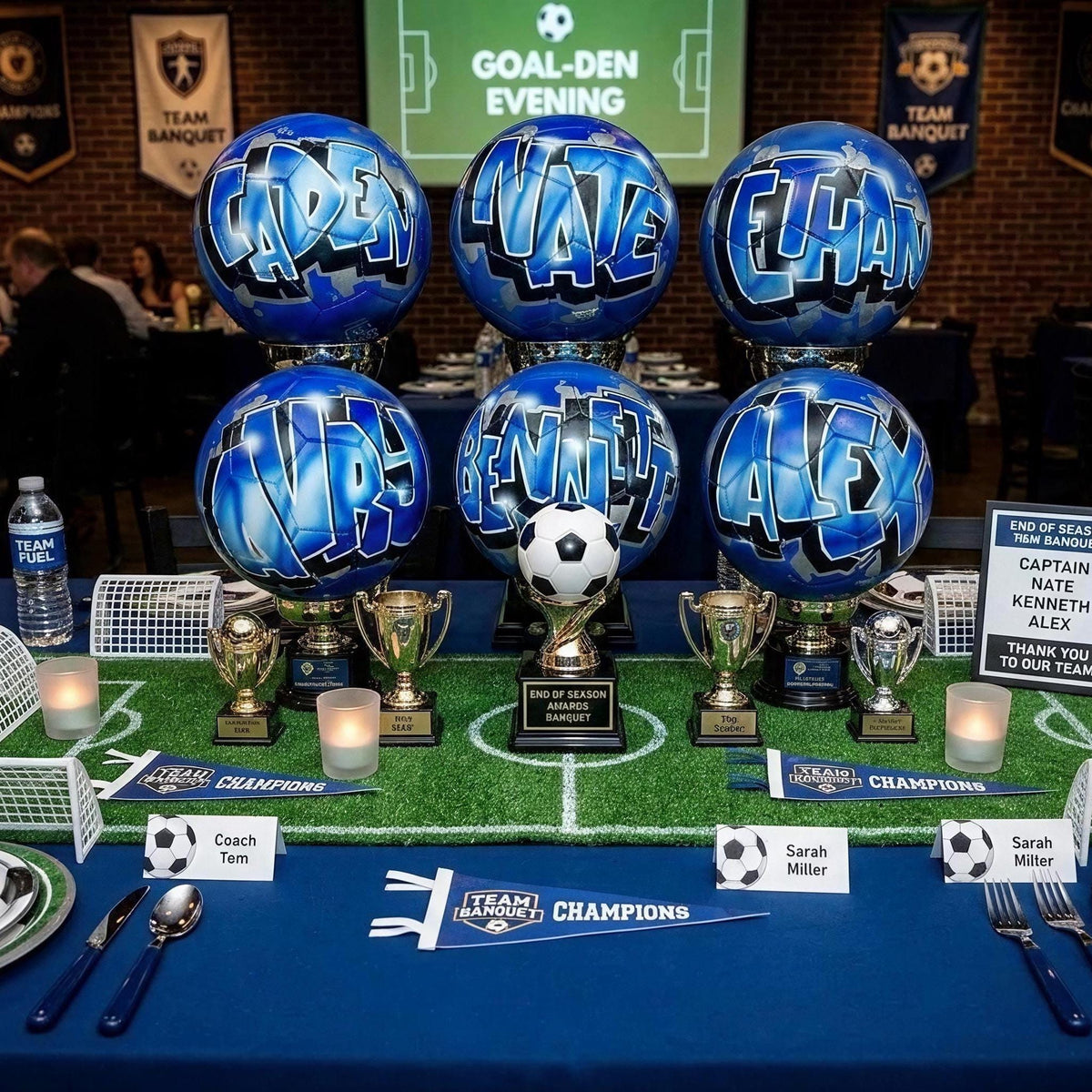 a table set up for a soccer-themed event, with various soccer-related items and decorations. There are several soccer balls, trophies, and banners arranged on the table, creating a festive atmosphere. The table also has plates, cutlery, and water bottles, indicating that it is prepared for a gathering or celebration.