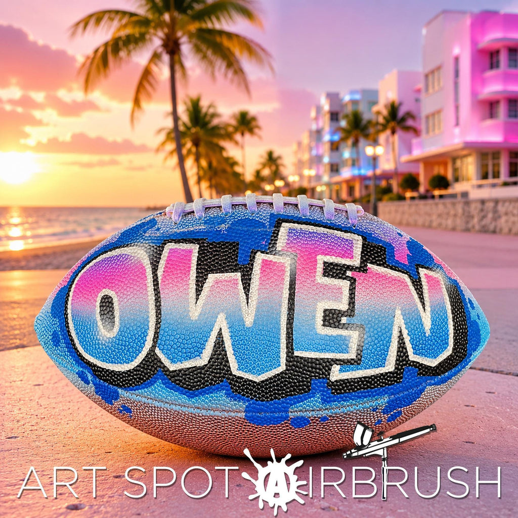 A custom football personalized with the name "OWEN" painted on it, placed on a beach at sunset with palm trees and buildings in the background.
