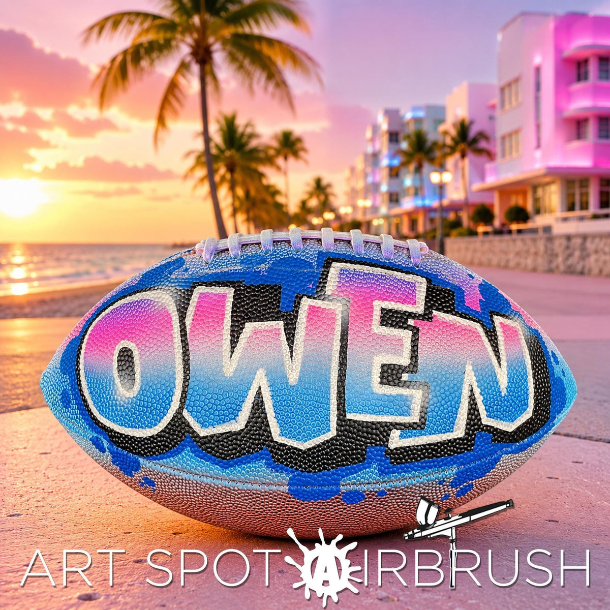 A custom football personalized with the name "OWEN" painted on it, placed on a beach at sunset with palm trees and buildings in the background.