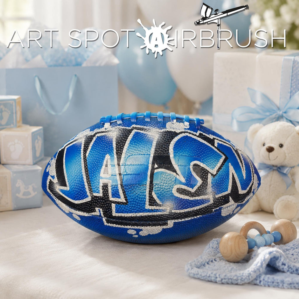 a blue personalized custom football with the name "JALEN" painted on it, surrounded by baby items such as a teddy bear and baby blocks, suggesting a baby shower or a child's room decoration.