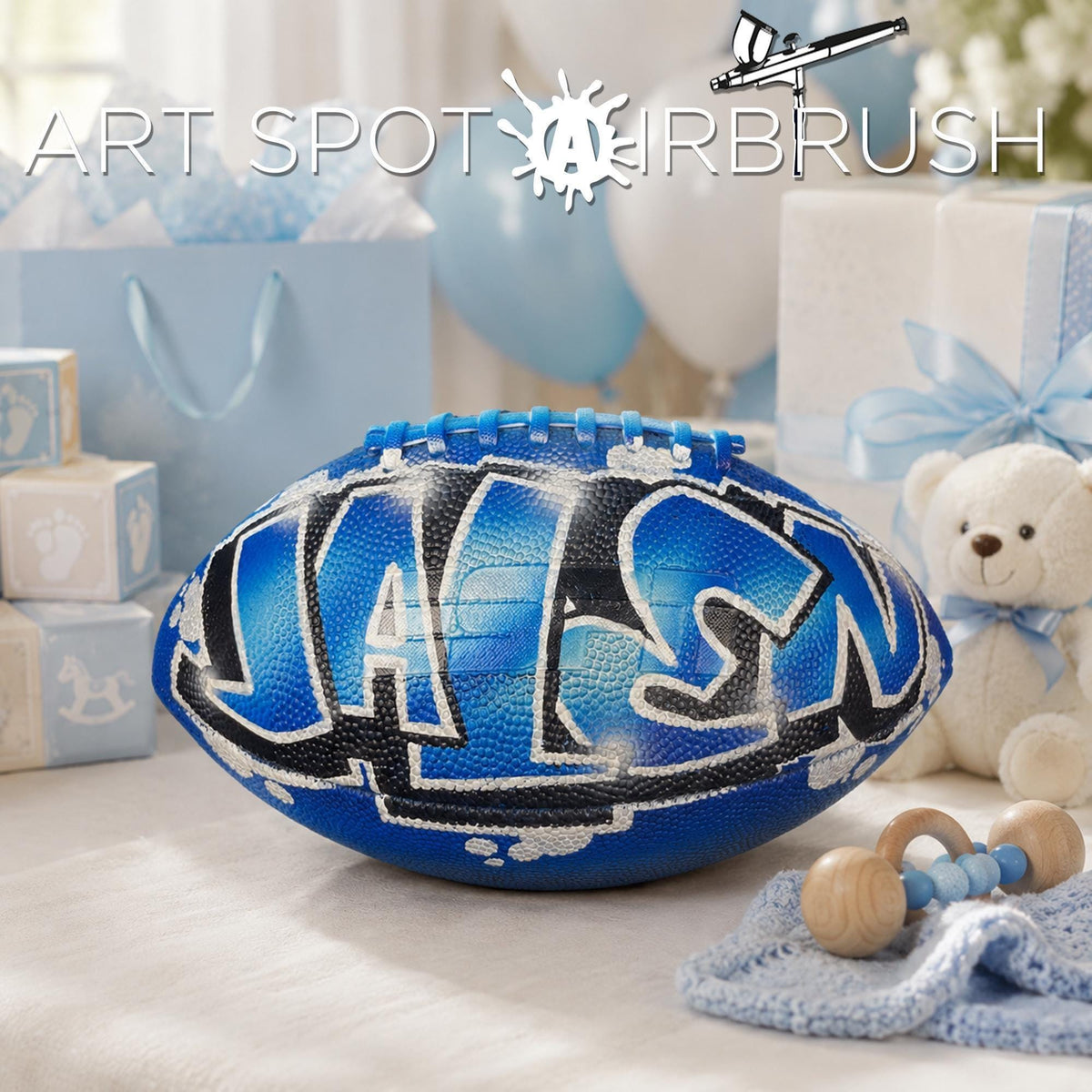a blue personalized custom football with the name "JALEN" painted on it, surrounded by baby items such as a teddy bear and baby blocks, suggesting a baby shower or a child's room decoration.