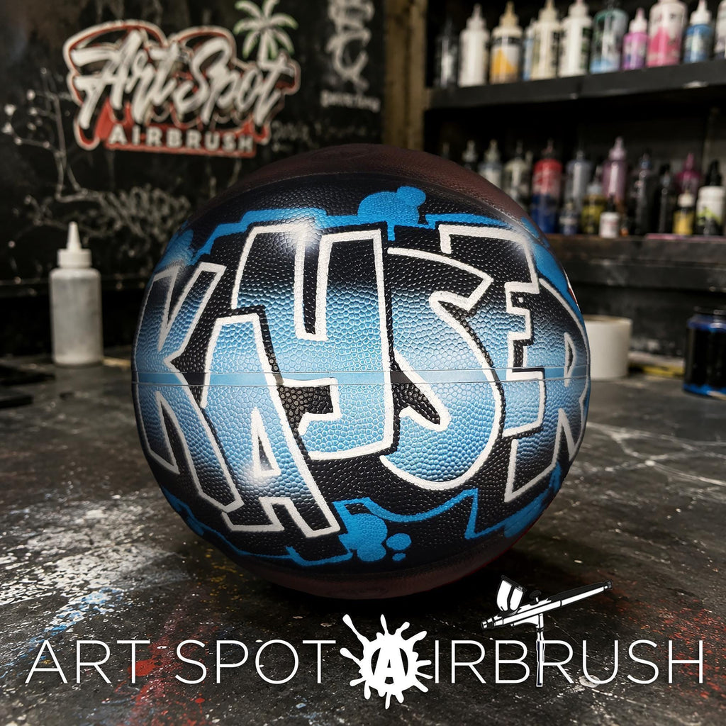 A basketball with a custom name painted on it, sitting on a table in front of a wall with graffiti art.