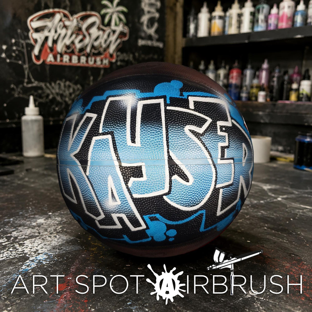 A basketball with a custom name painted on it, sitting on a table in front of a wall with graffiti art.