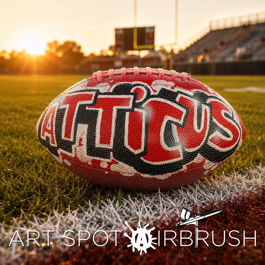 A custom football personalized with the word "ATTICUS" painted on it, resting on a grassy field with a scoreboard and stadium in the background during sunset.