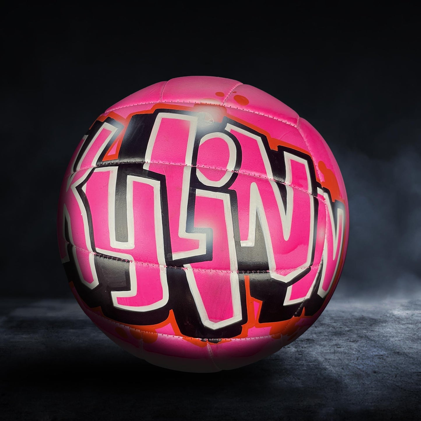 A pink and black soccer ball with graffiti-style text on it, placed on a dark surface.