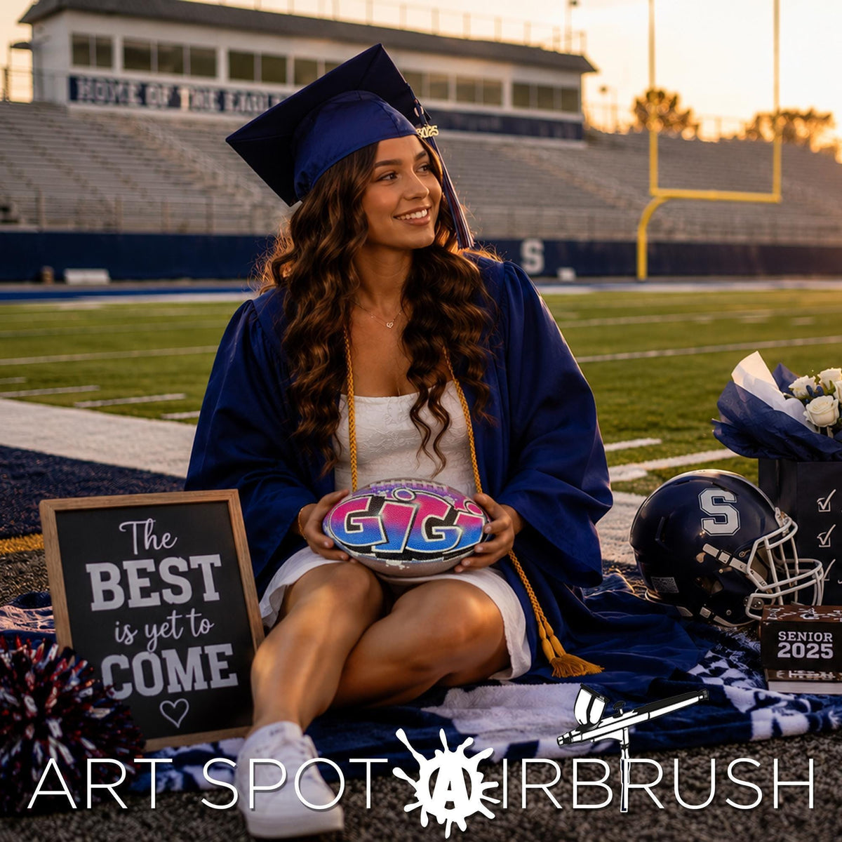 A smiling young woman in a graduation gown and cap sits on a blanket in front of a sign that says "The best is yet to come" and a football helmet, with a football field and stadium in the background. She is holding a Pink and Blue custom football, personalized with the name Gigi.