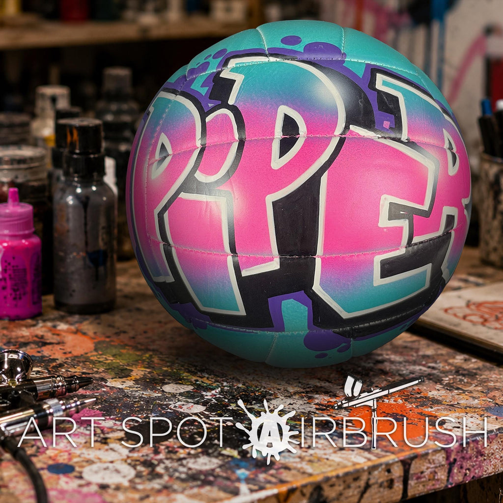A colorful, graffiti-style custom volleyball with the word "PIPER" painted on it, sitting on a artist's work surface with various art supplies and equipment.