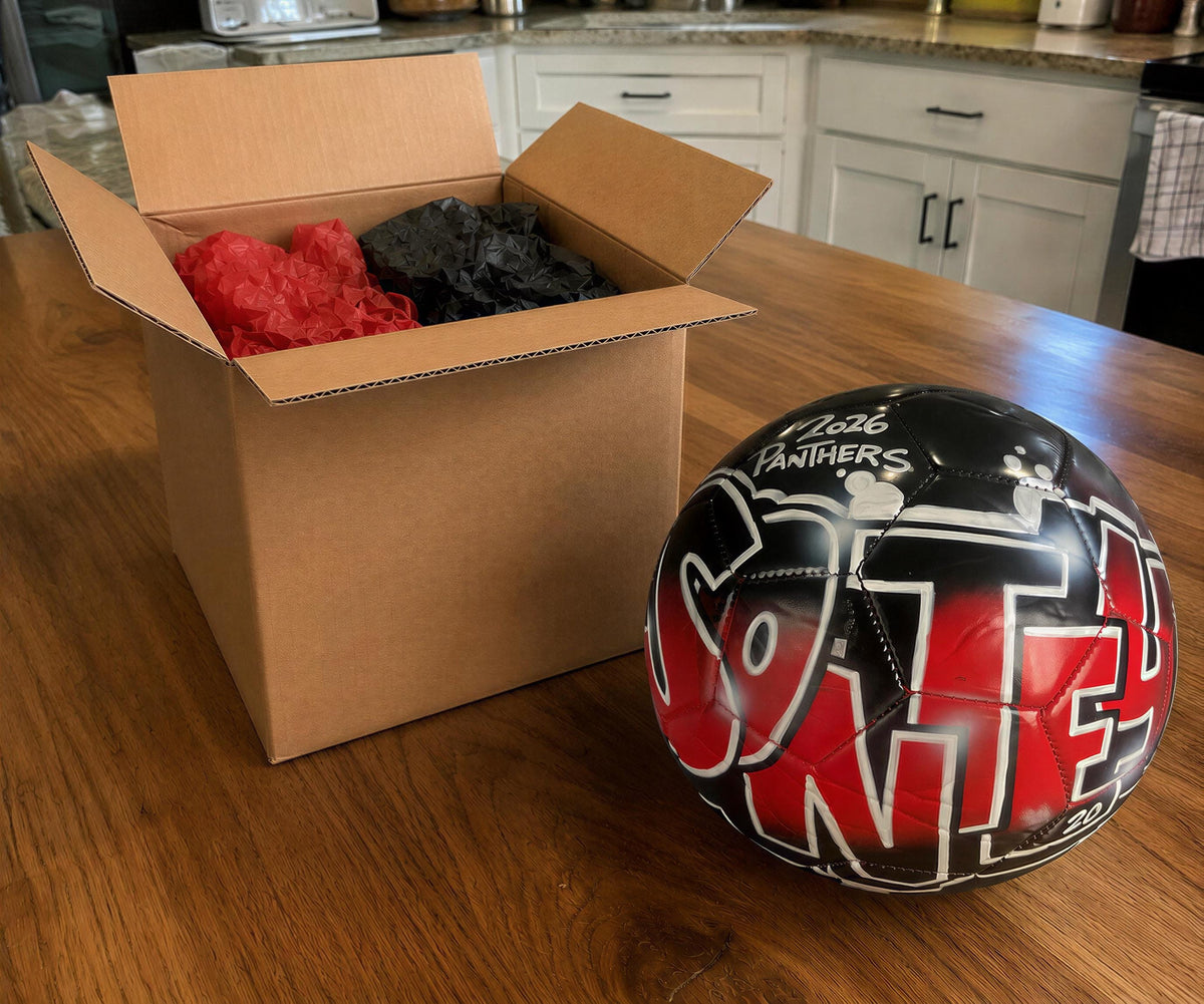 A cardboard box containing a red object and a black object is placed on a wooden table, next to a sports ball with graffiti on it.