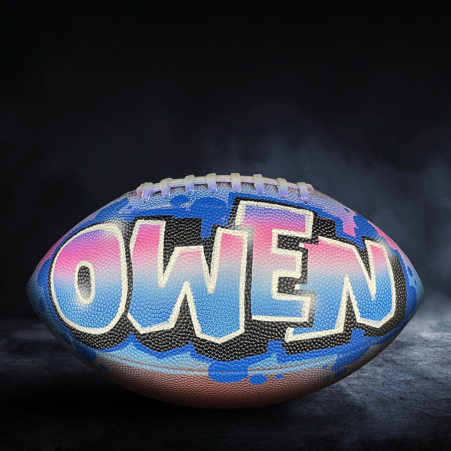 A football with the name "OWEN" painted on it, set against a dark background with smoke or fog.