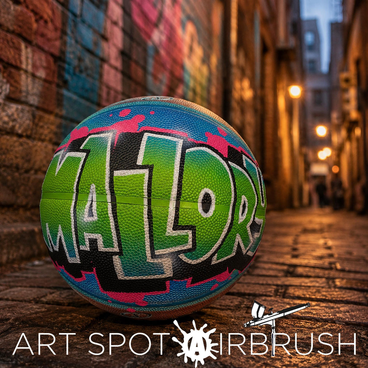 A colorful basketball with the word "MALOR" painted on it, sitting on a brick sidewalk in an urban setting with a graffiti-covered wall in the background.