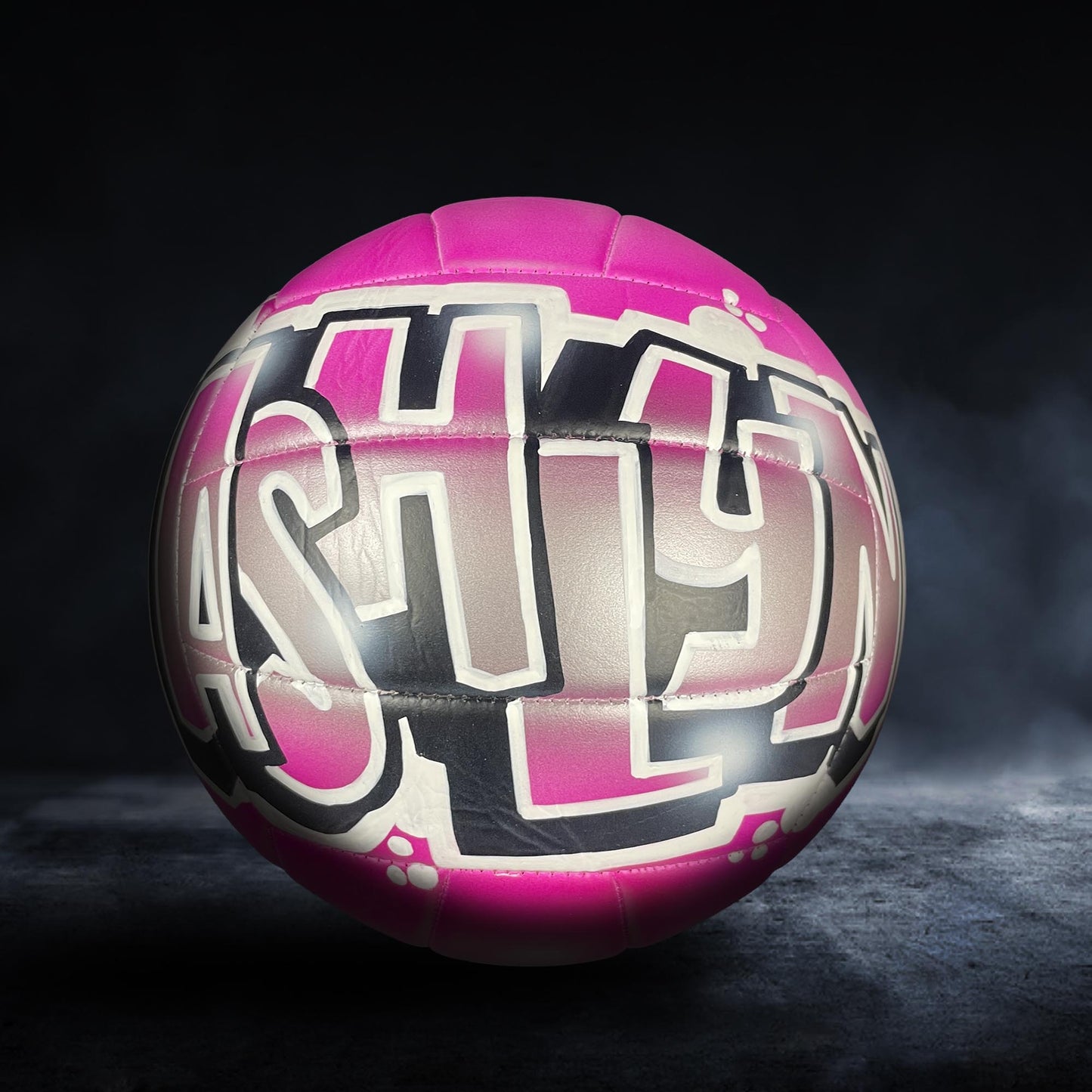 A pink and black volleyball with the word "Ashley" painted on it, against a dark background.