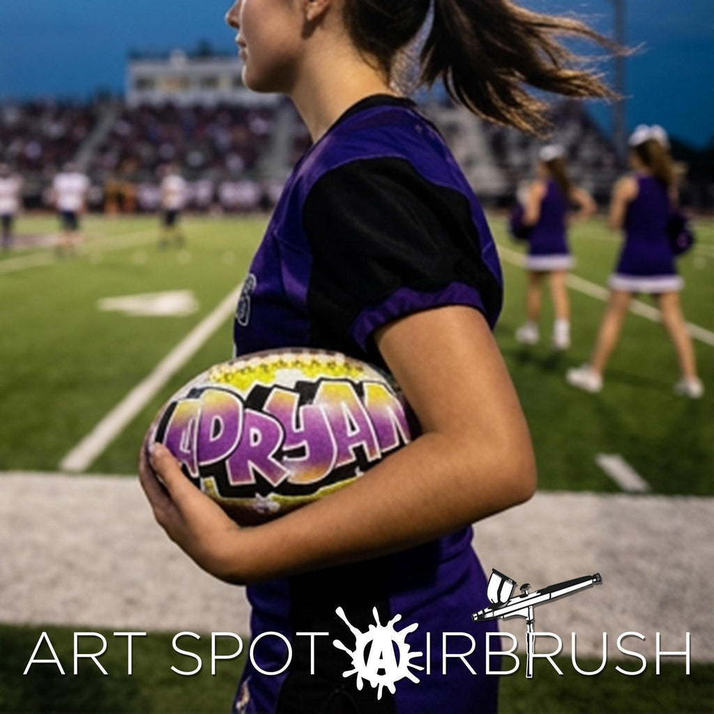 A young woman in a purple uniform is holding a colorful custom football personalized with the word "ADRYAN" written on it, standing on a football field.