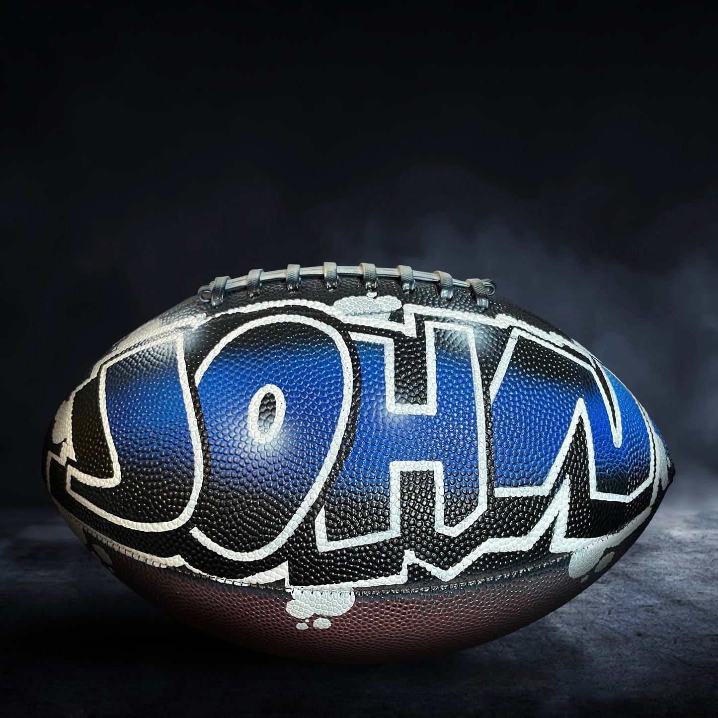 A football with the name "JOHN" painted on it, sitting on a dark surface with a misty background.