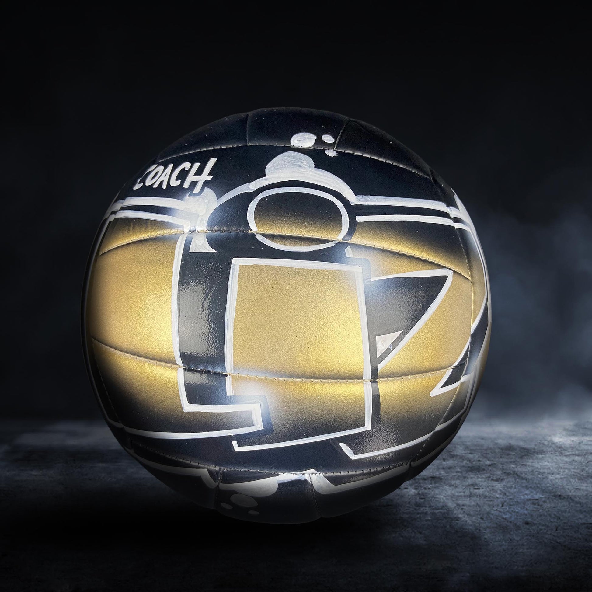 A black and gold volleyball with the word "COACH" written on it, sitting on a dark surface with a misty background.