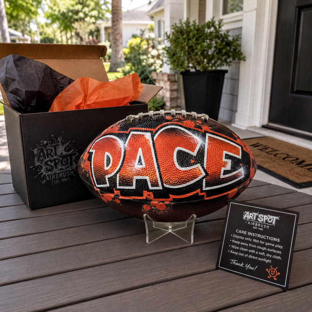 a custom football personalized with the word "PACE" painted on it, placed on a wooden surface next to a cardboard box and a small informational card.