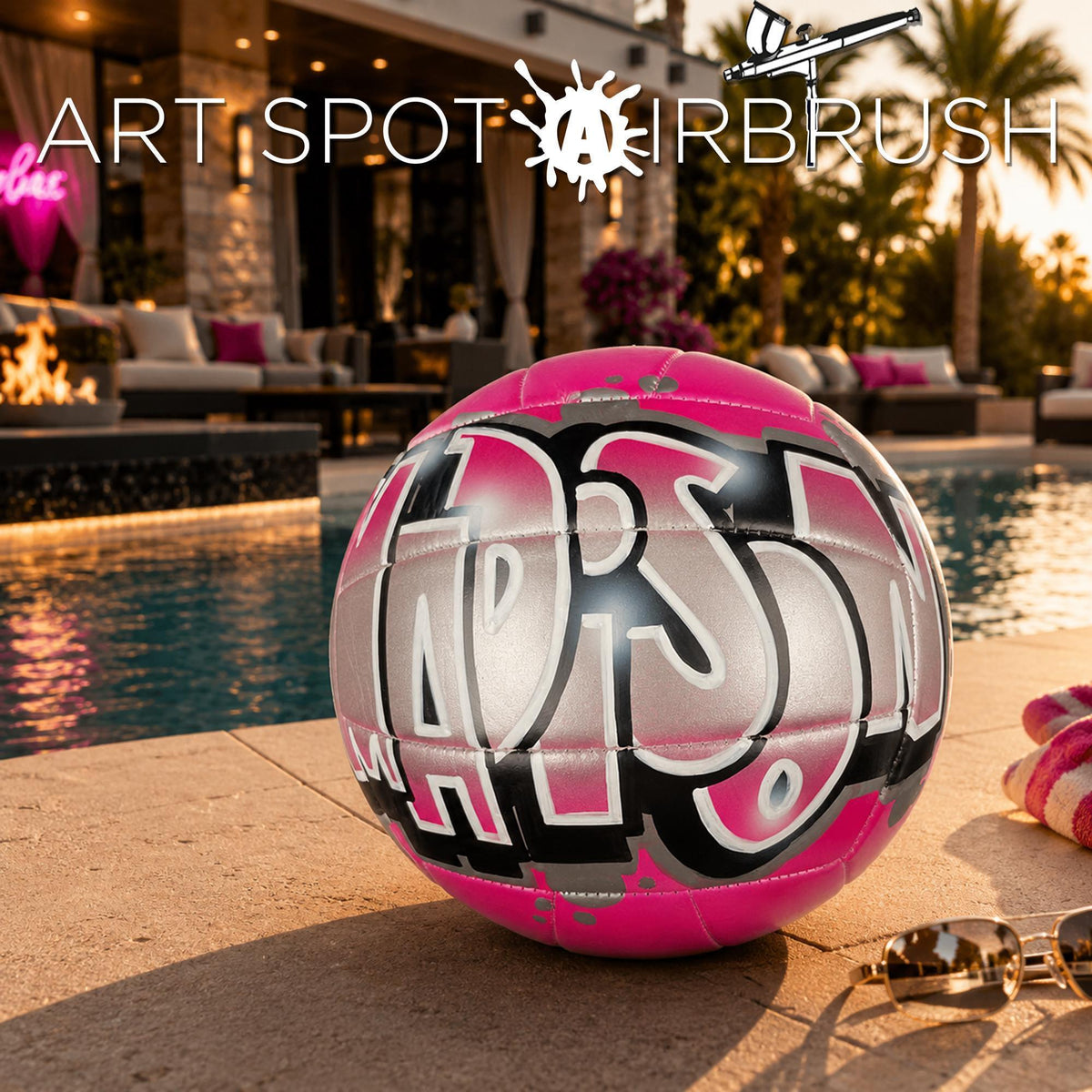 A pink and black custom volleyball personalized with the word "Madison" written on it, sitting on a tiled surface near a pool and palm trees.