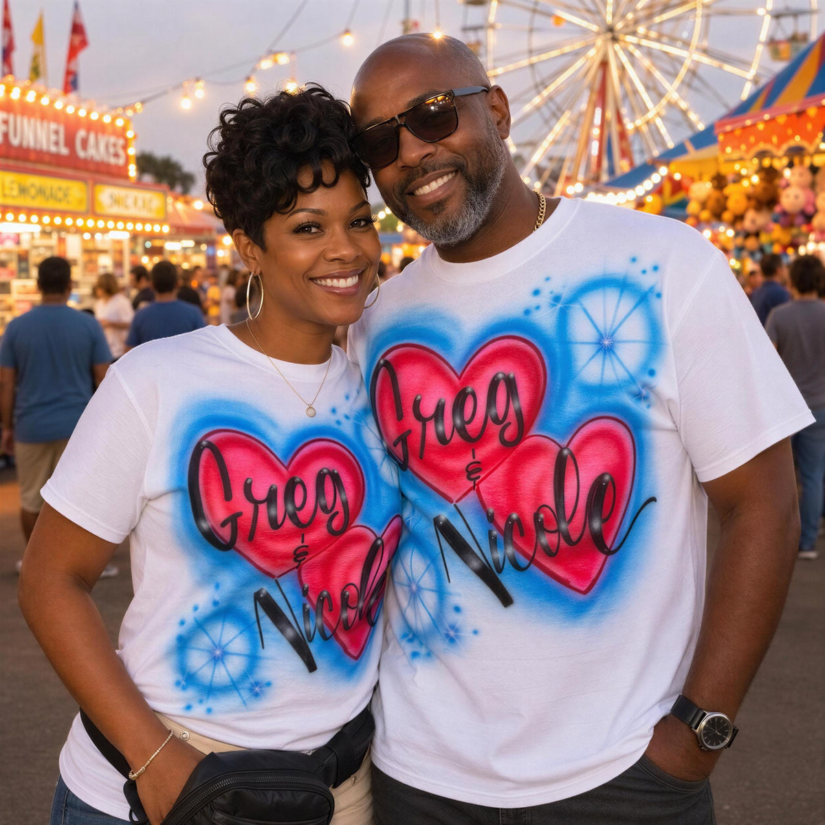 A smiling couple wearing matching t-shirts with a heart design stands together at an amusement park, with a ferris wheel and other carnival attractions visible in the background.