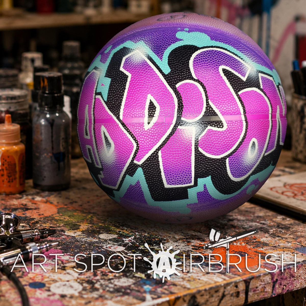 A colorful basketball with graffiti-style lettering is displayed on a cluttered work surface, surrounded by various art supplies and equipment.