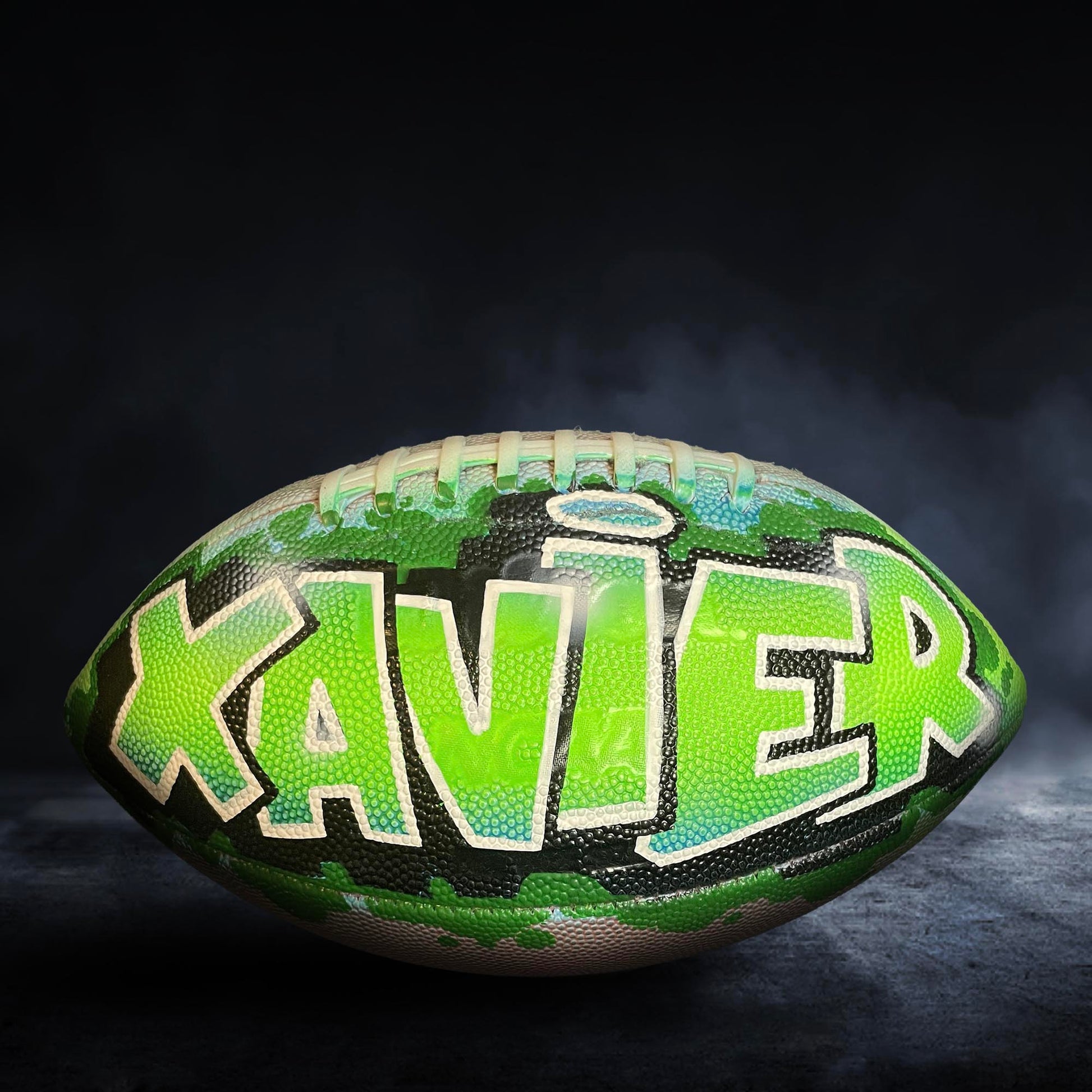A football with the name "XAVIER" painted on it, set against a dark background with smoke or fog.