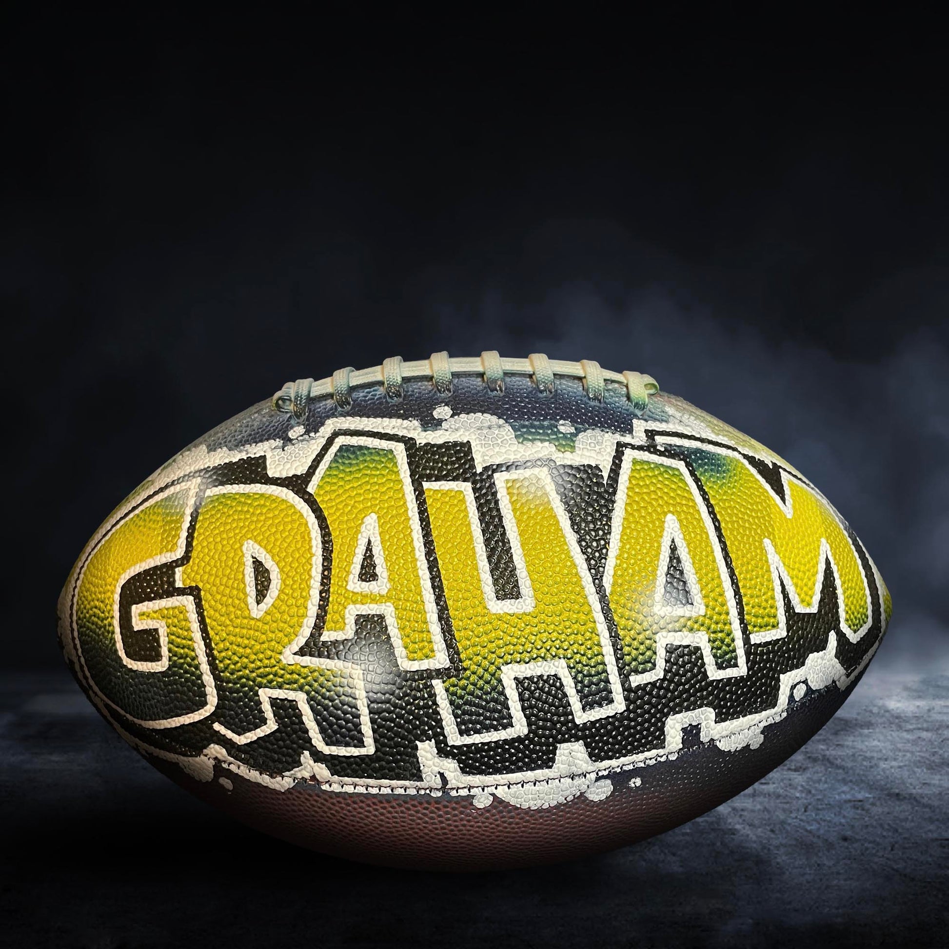 A football with the name "GRAHAM" painted on it, sitting on a dark surface with smoke or fog in the background.