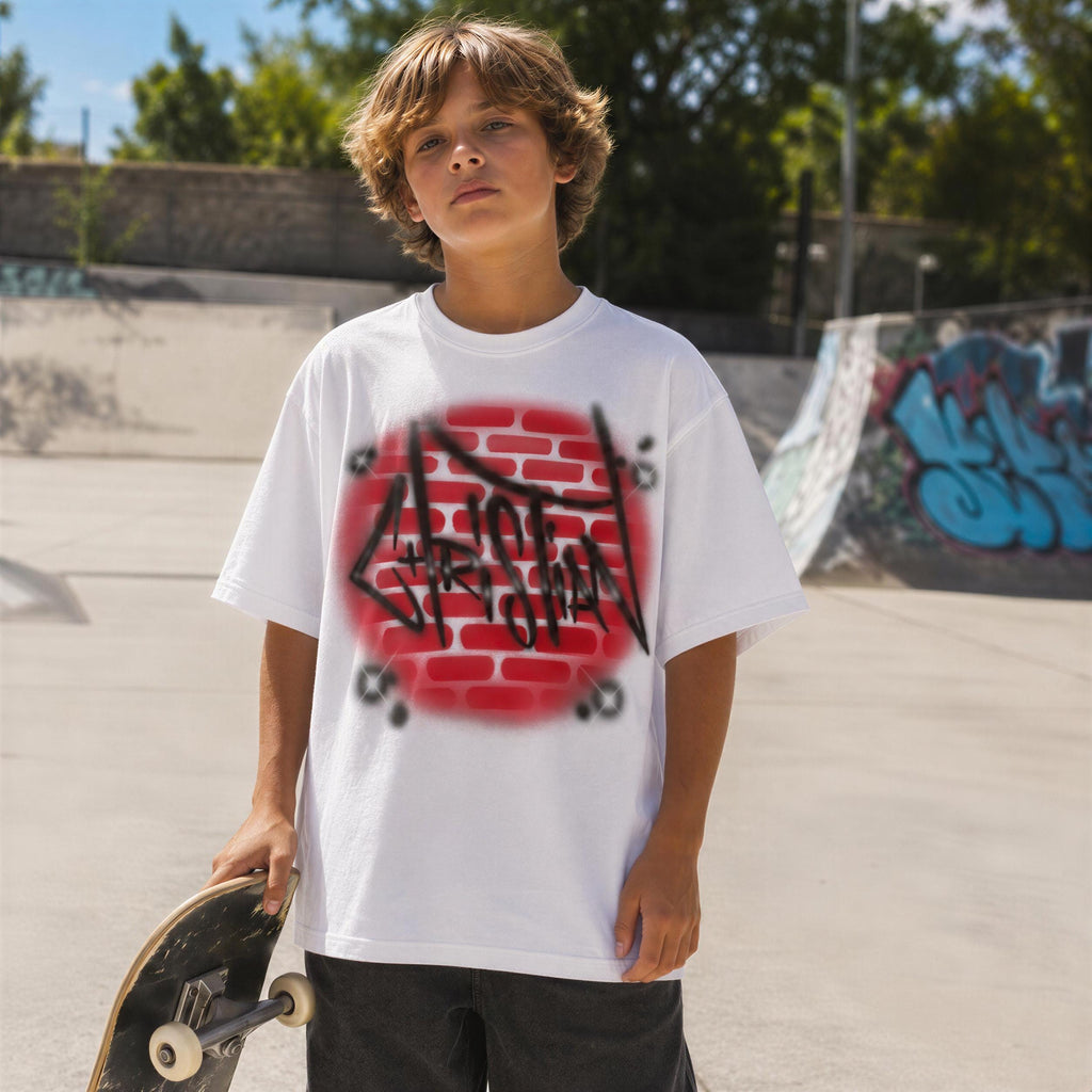 A young boy wearing a white t-shirt with graffiti on it, holding a skateboard, standing in front of a graffiti-covered ramp.