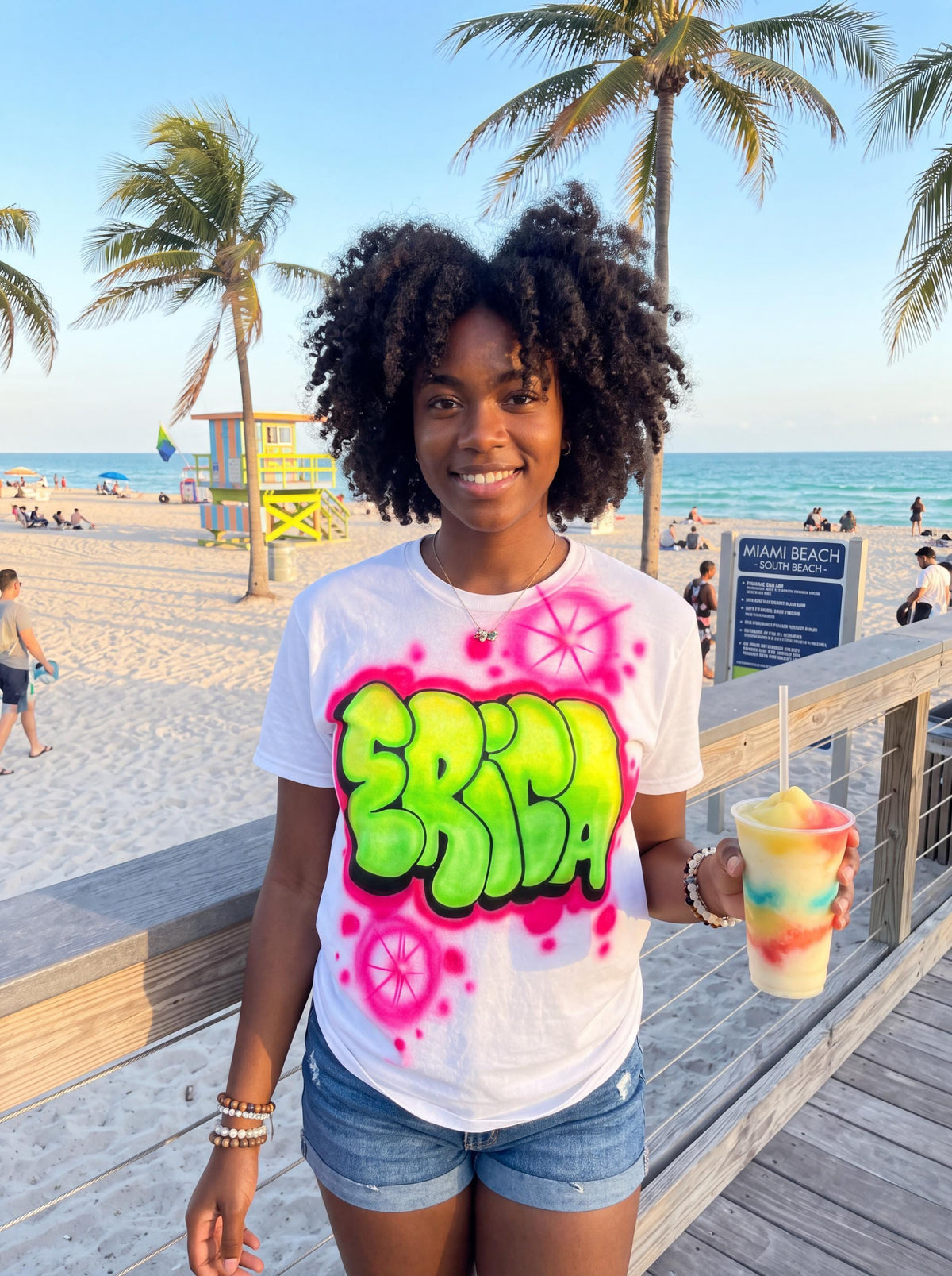 A young woman with curly hair stands on a boardwalk, holding a colorful drink and wearing a white t-shirt with a graffiti-style design.